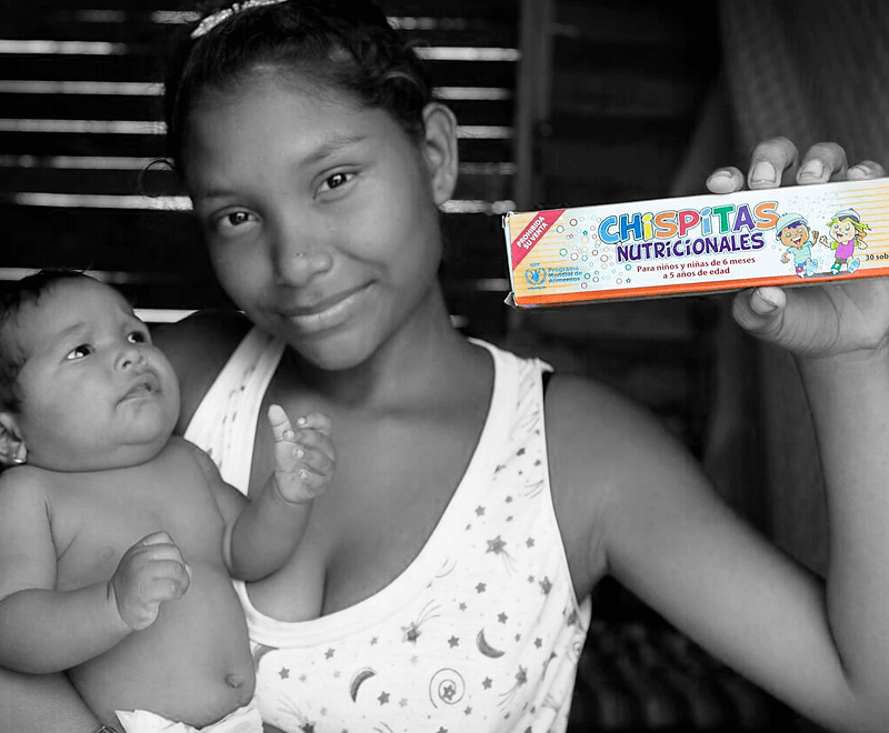 A woman holds her baby in one arm and a box of micronutrient powder, or "magical sprinkles," in the other