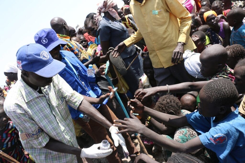 A WFP staffer is surrounded on all sides by people with children reaching out to him as he puts ink on their hands
