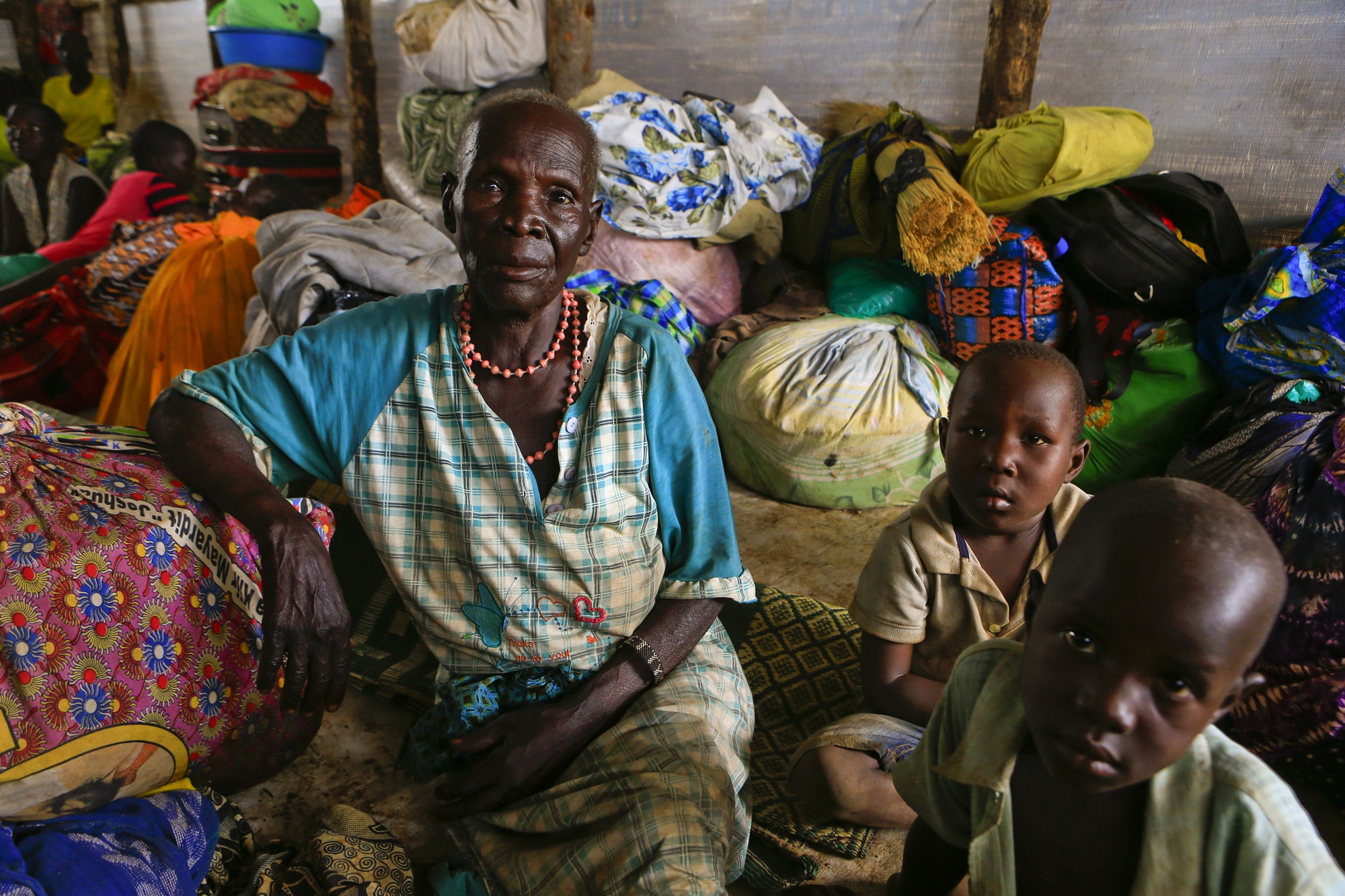 Abeer sits wearing a blue and white plaid dress alongside her two grandchildren in a tent filled with belongings of families who fled their homes in Pajok, South Sudan