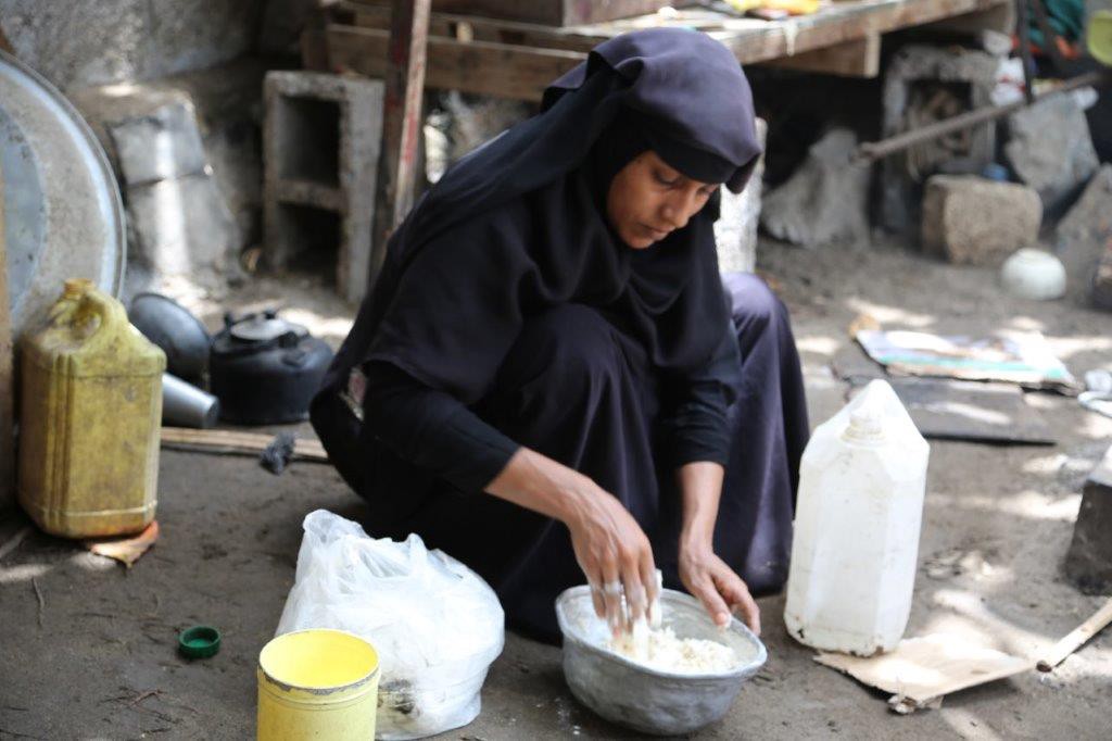 Aisha in her black hijab kneels on the ground and kneads bread in a tin bowl