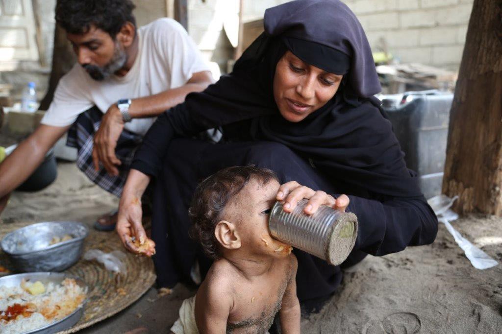 Aisha in her black hijab feeds Wafaa water out of a tin can, her husband Fadl sits behind them