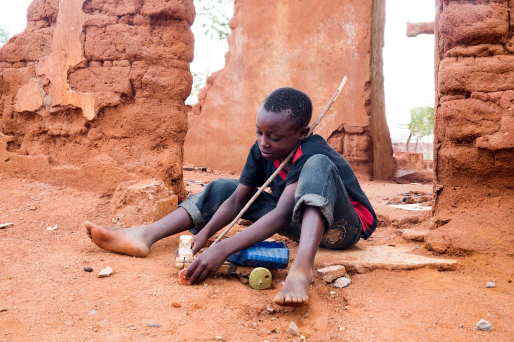 A boy sits on the ground with a racing car he made out of sticks, string and an empty water bottle.