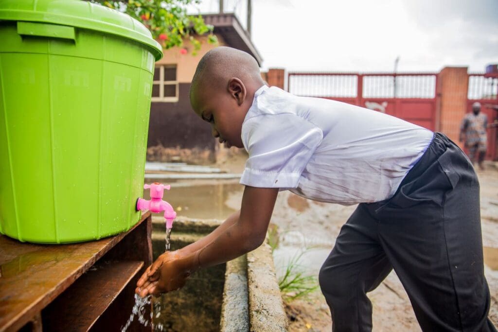 A young boy in a school uniform leans over to wash his hands at a hand washing station.