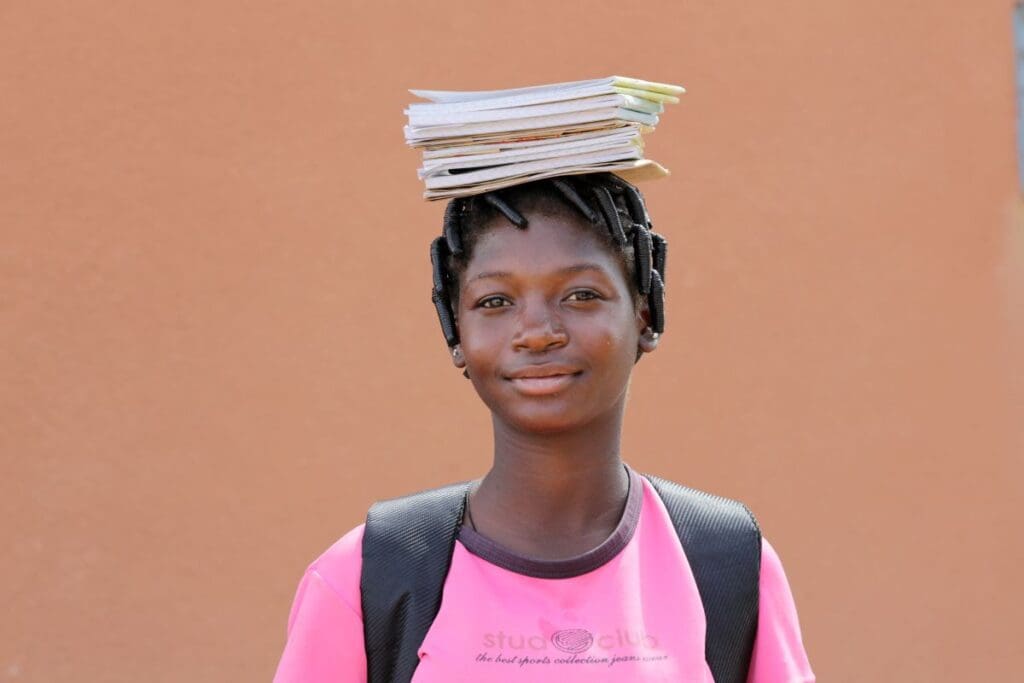 A girl in a pink shirt carries a stack of papers on her head while looking at the camera.