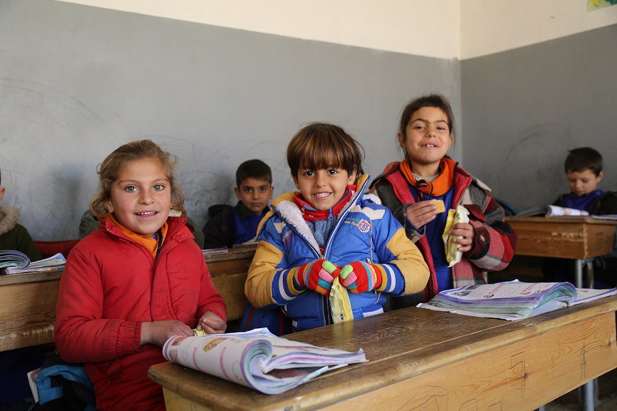 Three girls in colorful winter coats stand at a desk in a school classroom