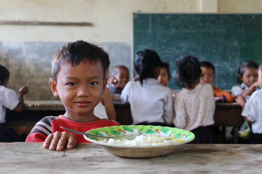 A schoolboy sits at a table in front of a bowl of food, looking at the camera. Other students sit behind him.