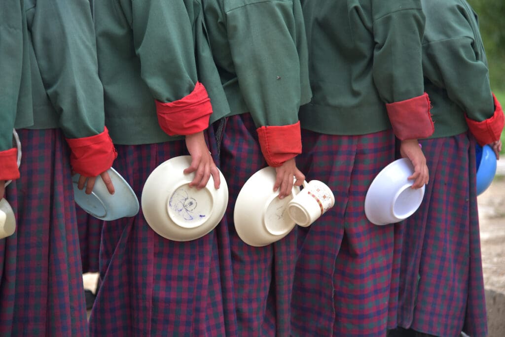 Students stand in a line holding empty food bowls, only their arms are pictured.