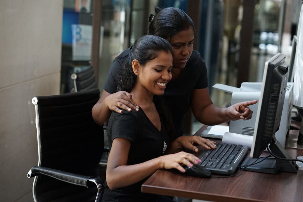 Two young women in black t-shirts work together at a computer.