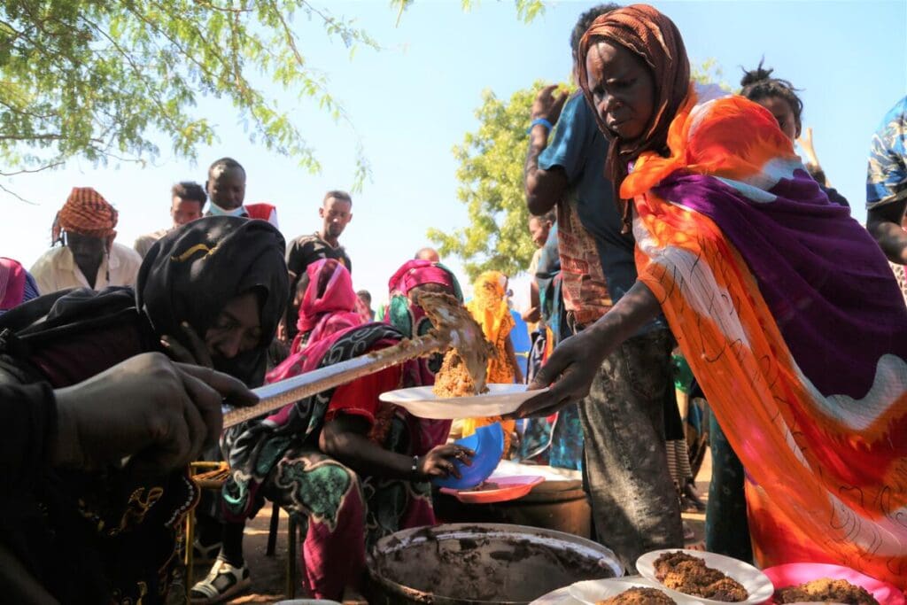 A woman holds out a plate, and volunteers serve her food from plates on the ground.