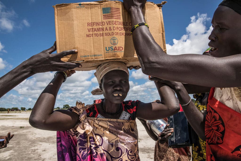 A woman holds a box of food on her head, with arms and hands reaching into the photo to help her.