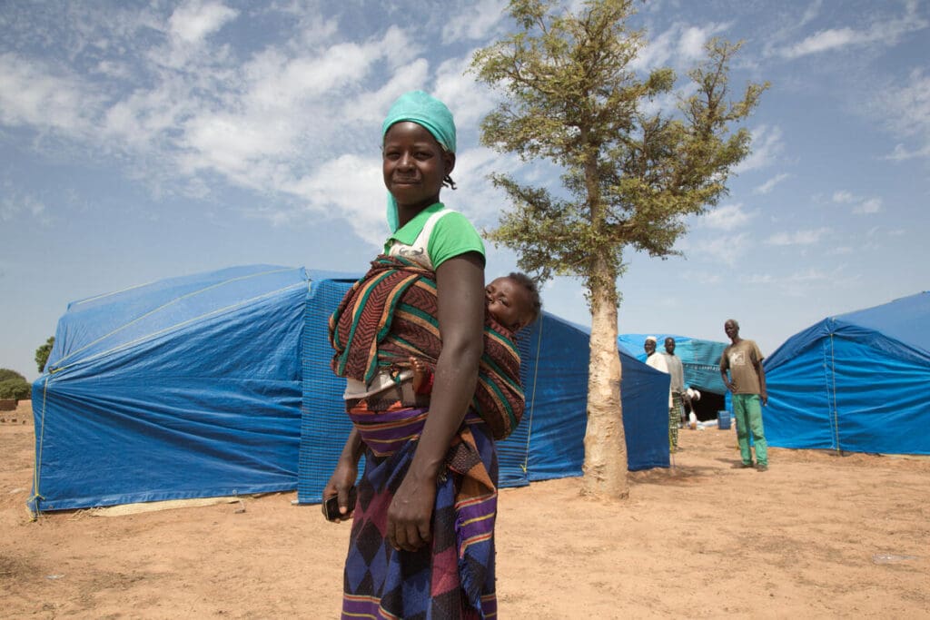 A woman stands outside of a refugee tent, her baby wrapped around her back with a cloth.