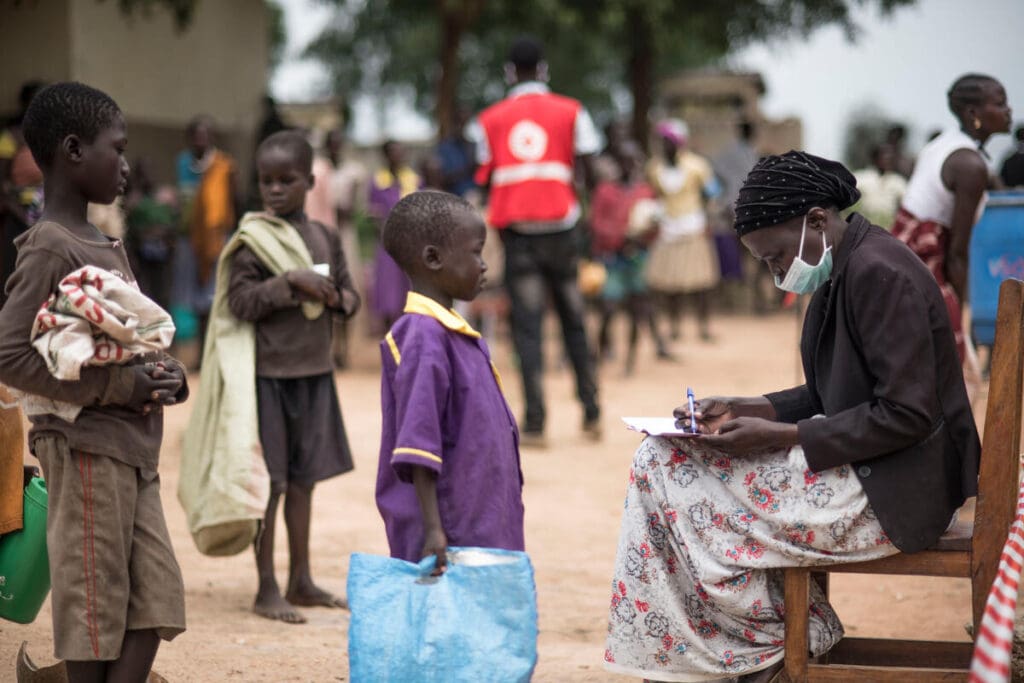 Schoolkids line up in front of a teacher sitting in a chair with a clipboard.