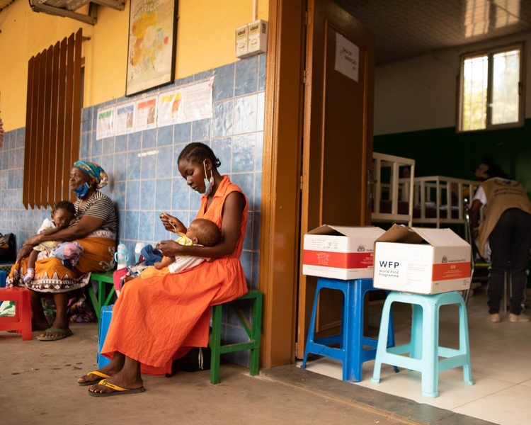 A woman in an orange dress sits on a blue chair, holding a baby.