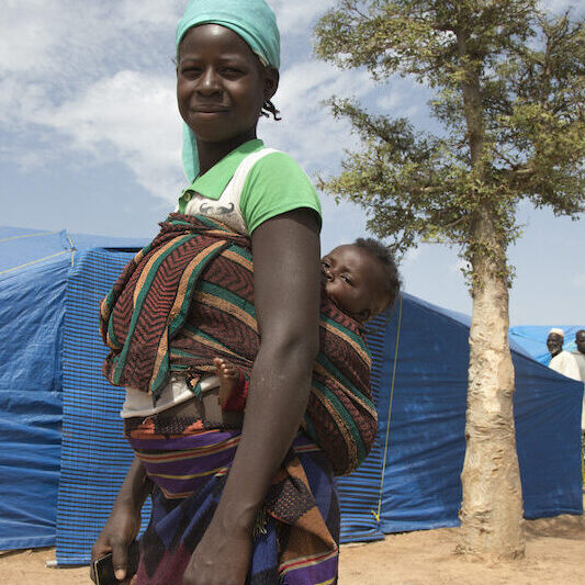 A woman stands in front of a blue tent, a baby sleeping on her back.