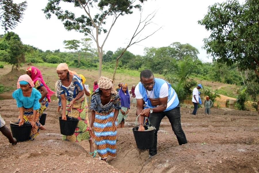 A UN volunteer in a blue vest works farmland with a group of women.